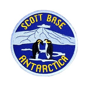 May include: A blue and white embroidered patch with the text "Scott Base Antarctica" in yellow lettering. Two penguins are standing on a white ice shelf in front of a mountain with a white peak.