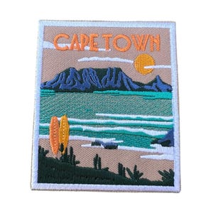 Puede incluir: Parche bordado con un paisaje de Ciudad del Cabo y el texto "CAPE TOWN". El diseño incluye una cordillera, el océano, el sol y dos tablas de surf. El parche tiene un fondo beige con un borde blanco.