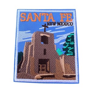 Può includere: Toppa ricamata con sfondo blu e bordo bianco. La toppa presenta un edificio marrone con una croce in cima e il testo "SANTA FE NEW MEXICO" in arancione.