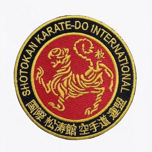 Könnte beinhalten: Ein kreisförmiger gestickter Aufnäher mit roter Mitte und einer gelben Tigerillustration. Der schwarze Rand enthält den gelben Text SHOTOKAN KARATE-DO INTERNATIONAL sowie japanische Schriftzeichen. Das Design hat einen gelben Außenrand.