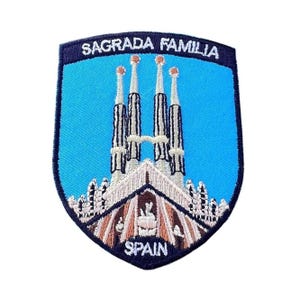 May include: A blue and white embroidered patch with the text "Sagrada Familia" and "Spain". The patch features a depiction of the Sagrada Familia basilica in Barcelona, Spain.