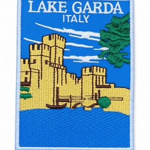 Könnte beinhalten: Ein rechteckiger gestickter Aufnäher mit einer gelben Burg mit Türmen auf blauem Grund. Der Text LAKE GARDA ITALY ist oben in Weiß gestickt. Ein grüner Baum befindet sich auf der rechten Seite und ein kleines braunes Boot liegt auf dem blauen Wasser.
