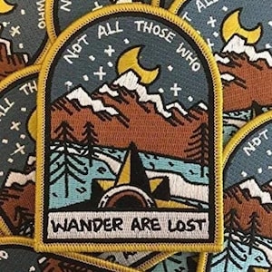 Może przedstawiać: Naszywka haftowana na żółto i niebiesko z górskim krajobrazem i rosnącym księżycem. Tekst brzmi "Not all those who wander are lost."