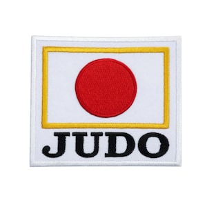 Pode incluir: Um patch quadrado com o design da bandeira japonesa: um círculo vermelho sobre fundo branco, emoldurado por uma borda amarela. A palavra "JUDO" é bordada em letras pretas abaixo.