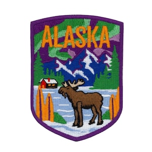 Op de afbeelding: Geborduurde patch met het woord "ALASKA" in oranje letters. Het ontwerp toont een eland, bergen, bomen en een hut, tegen een paarse en groene achtergrond. Paarse rand.