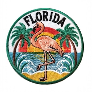 Puede incluir: Parche bordado con un flamenco rosa en una playa, palmeras y una puesta de sol. La palabra "FLORIDA" está arqueada en letras negras, dentro de un borde verde. Diseño de temática tropical.