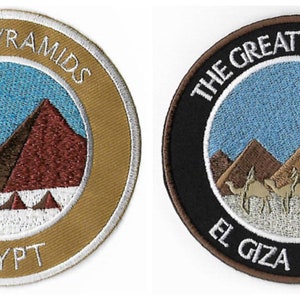 Könnte beinhalten: Zwei gestickte Aufnäher, die die großen Pyramiden von Gizeh in Ägypten darstellen. Der erste Aufnäher ist goldfarben mit weißem Text und zeigt die Pyramiden vor einem blauen Himmel. Der zweite Aufnäher ist schwarz mit braunem Text und zeigt die Pyramiden vor einem blauen Himmel mit einer Sonne und Kamelen im Vordergrund.
