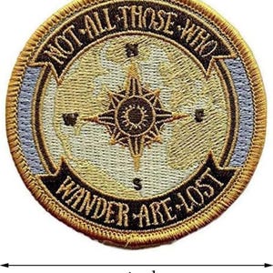 Puede incluir: Parche bordado circular con un diseño de rosa de los vientos y el texto "NOT ALL THOSE WHO WANDER ARE LOST". El parche mide aproximadamente 7,6 cm de diámetro, con una combinación de colores dorados, azules y marrones. El diseño tiene una apariencia detallada y texturizada.