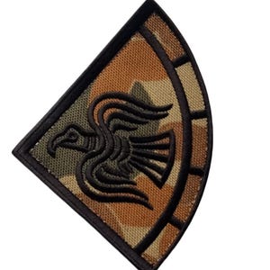 May include: Embroidered patch featuring a stylized black bird design on a camouflage background. The patch has a black border and a curved, segmented design on the right side. The color palette includes shades of brown, green, and black.