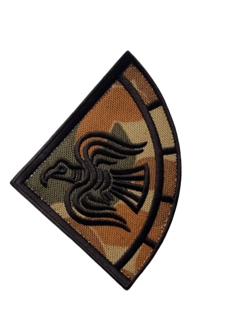 Op de afbeelding: Geborduurde patch met een gestileerd zwart vogelontwerp op een camouflage achtergrond. De patch heeft een zwarte rand en een gebogen, gesegmenteerd ontwerp aan de rechterkant. De kleuren zijn bruin, groen en zwart.