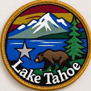 Könnte beinhalten: Gestickter Aufnäher mit einem Lake Tahoe-Design. Der Aufnäher zeigt eine Bergkette, einen See, einen Bären, einen Stern und eine Kiefer, mit den Worten "Lake Tahoe" in Weiß. Er hat einen goldenen Rand.