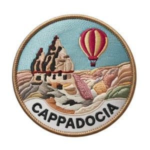 Pode incluir: Patch bordado com uma vista da Capadócia, Turquia. O design inclui um balão de ar quente, formações rochosas e a palavra "CAPPADOCIA". O patch tem um fundo azul claro e uma borda dourada.