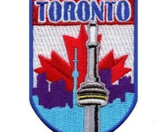 Puede incluir: Parche bordado con la palabra "TORONTO" en azul sobre un fondo rojo y blanco. El diseño incluye una hoja de arce roja, la Torre CN y un paisaje urbano en morado y azul. El parche tiene un borde azul.
