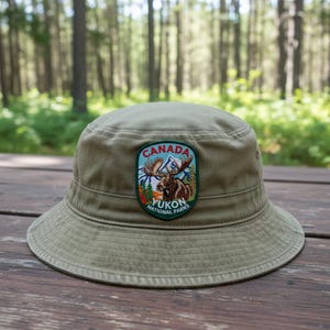 May include: An olive green bucket hat featuring a central embroidered patch. The patch displays a brown moose, blue mountains, green pine trees, and the text "CANADA YUKON NATIONAL PARKS" in white and red lettering on a dark green background.