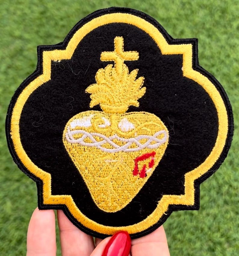 May include: A gold embroidered patch with a black background featuring a stylized heart with a cross and flames. The heart is surrounded by a white rope-like design and has a red drop of blood on the side.