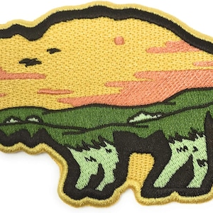 Op de afbeelding: Een geborduurde strijk-op patch in de vorm van een bizonsilhouet. De bison is omlijnd met zwart en gevuld met een gele achtergrond. De bovenkant van de patch heeft een zonsondergang met roze en oranje wolken. De onderkant van de patch toont een groen grasveld met een paar kleine heuvels.