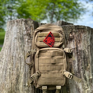 May include: A tan tactical backpack with a black and red patch featuring a bird design. The backpack has multiple straps and buckles.