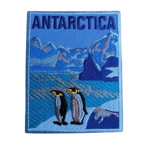 Puede incluir: Parche bordado con un paisaje antártico azul y blanco con la palabra "ANTARCTICA" en la parte superior. Dos pingüinos están de pie sobre una superficie nevada. El parche tiene un borde azul.