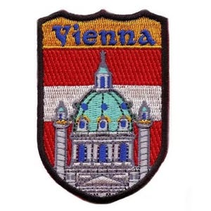 May include: An embroidered patch with a shield shape, featuring the word "Vienna" in yellow lettering on a gold background. The shield also depicts a white and red Austrian flag with a detailed illustration of a building with a green dome.
