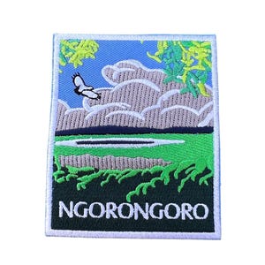 Può includere: Toppa ricamata con una scena di paesaggio verde, blu e bianco. La toppa ha un bordo bianco e il testo "NGORONGORO" in lettere bianche su sfondo verde.