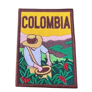 May include: Embroidered patch featuring a cartoon illustration of a person wearing a hat and picking coffee beans in a field. The patch has a brown border and the word "COLOMBIA" in brown text on a yellow background.