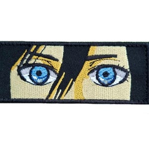 Könnte beinhalten: Ein schwarzer und beiger gestickter Aufnäher mit einem Paar blauer Augen. Die Augen sind von einem schwarzen Hintergrund und einem beigen Gesicht umgeben.