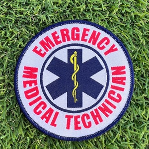Op de afbeelding: Een ronde witte patch met blauwe en rode rand. In het midden van de patch bevindt zich een blauwe ster van het leven met een gele slang en staf. De tekst "EMERGENCY MEDICAL TECHNICIAN" staat in rood rond de ster van het leven.