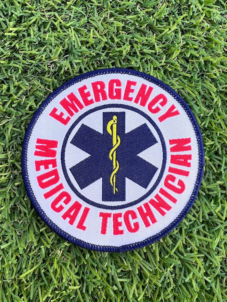 Op de afbeelding: Een ronde witte patch met blauwe en rode rand. In het midden van de patch bevindt zich een blauwe ster van het leven met een gele slang en staf. De tekst "EMERGENCY MEDICAL TECHNICIAN" staat in rood rond de ster van het leven.