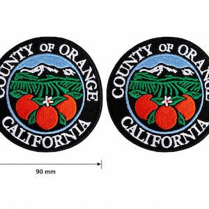 May include: Two round black patches with the words "COUNTY OF ORANGE CALIFORNIA" in white. The patches feature a design of oranges, green fields, and mountains. Each patch is 90 mm in diameter.