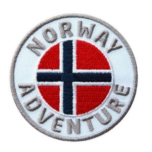 Può includere: Un cerotto circolare bianco con un bordo grigio. Il centro del cerotto presenta la bandiera norvegese in rosso, bianco e blu. Le parole "NORWAY ADVENTURE" sono scritte in un carattere circolare grigio attorno alla bandiera.