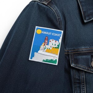 Peut inclure: Veste en jean bleu fonc&eacute; avec un &eacute;cusson rectangulaire repr&eacute;sentant un paysage c&ocirc;tier. L'&eacute;cusson affiche un soleil jaune, un ciel bleu et des b&acirc;timents blancs avec le texte "AMALFI COAST". La veste a un devant boutonn&eacute; et une poche poitrine.