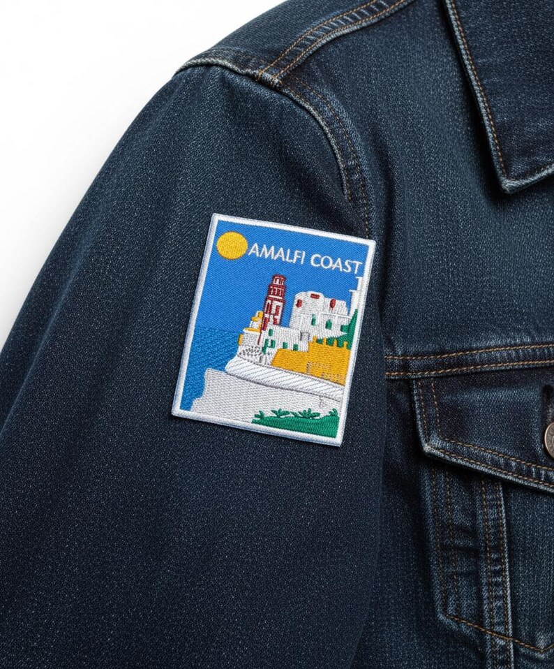 Peut inclure: Veste en jean bleu fonc&eacute; avec un &eacute;cusson rectangulaire repr&eacute;sentant un paysage c&ocirc;tier. L'&eacute;cusson affiche un soleil jaune, un ciel bleu et des b&acirc;timents blancs avec le texte "AMALFI COAST". La veste a un devant boutonn&eacute; et une poche poitrine.