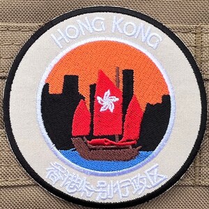 Może przedstawiać: Okrągła naszywka haftowana w kolorze białym i czarnym z napisem "Hong Kong" w białym kolorze wokół krawędzi. Środek naszywki przedstawia czerwony i biały żaglowiec z czerwonym zachodem słońca w tle. Tekst "香港特別行政區" jest napisany białym kolorem wokół dolnej części naszywki.