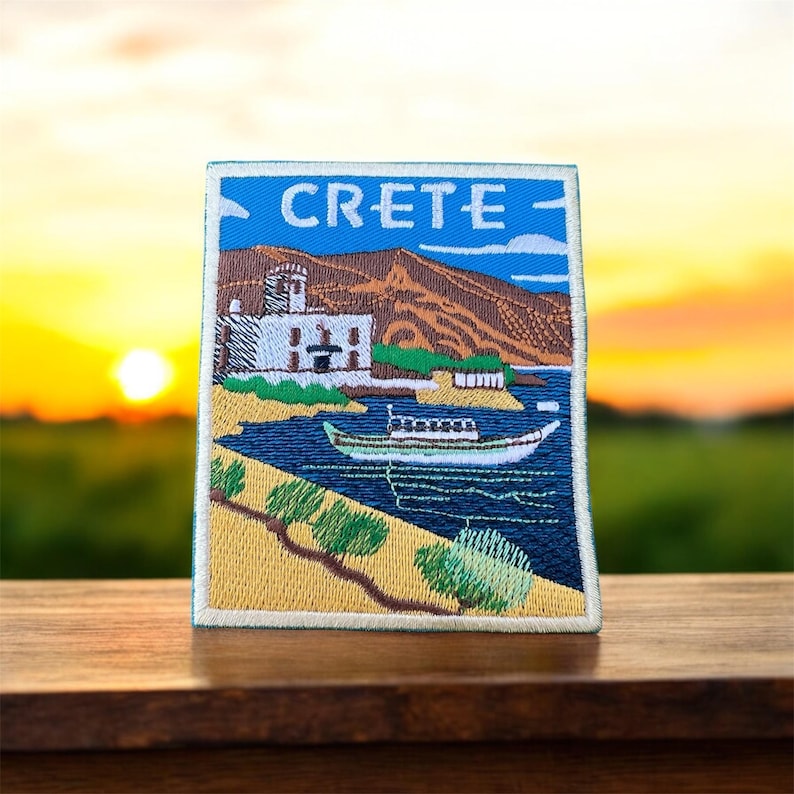 Peut inclure: Patch brod&eacute; avec une image color&eacute;e d'une petite ville sur la c&ocirc;te de Cr&egrave;te, en Gr&egrave;ce. Le patch repr&eacute;sente un b&acirc;timent blanc avec un toit bleu, un petit bateau dans l'eau et une colline verte en arri&egrave;re-plan. Le texte "CRETE" est brod&eacute; en blanc sur un fond bleu en haut du patch.