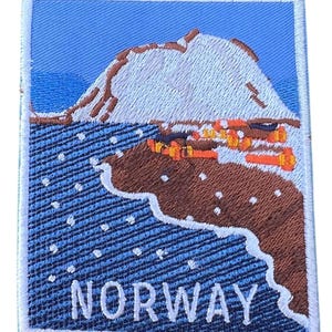 Könnte beinhalten: Gestickter Aufnäher mit einem weißen Berg, braunem Land und orangenen Gebäuden vor blauem Himmel mit weißen Schneeflocken. Der Text "NORWAY" ist in Weiß am unteren Rand des Aufnähers gestickt.