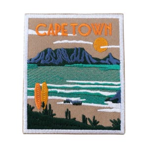 Könnte beinhalten: Gestickter Aufnäher mit einer Landschaft von Kapstadt. Das Design zeigt eine Bergkette, das Meer, die Sonne und den Text "CAPE TOWN" in Orange. Zwei orangefarbene Surfbretter sind am Strand. Der Aufnäher hat einen beigen Hintergrund und einen weißen Rand.