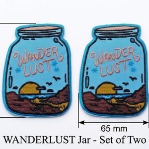 Könnte beinhalten: Zwei gestickte Aufnäher in Form von Gläsern, jeweils 90 mm hoch und 65 mm breit. Die Aufnäher sind hellblau mit einer Sonnenuntergangsszene und den Worten "Wanderlust". Die Aufnäher haben ein braunes, gelbes und blaues Design. Der Text "WANDERLUST Jar - Set of Two" steht darunter.