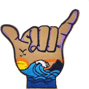 Op de afbeelding: Een geborduurde patch van een bruine hand die het shaka-teken maakt, met een zonsondergang, oceaan golven en palmbomen op de achtergrond.