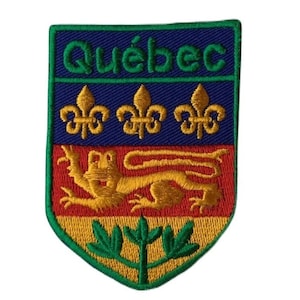 May include: An embroidered patch featuring the coat of arms of Quebec, Canada. The patch is shaped like a shield and features a blue background with three gold fleurs-de-lis, a red and gold lion, and a green plant with three leaves. The words "Quebec" are embroidered in green on a blue background at the top of the patch.
