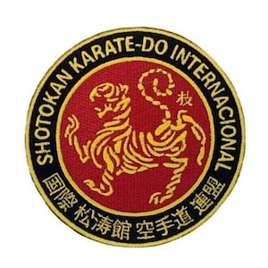 Può includere: Una toppa rotonda ricamata in oro con un bordo nero. La toppa presenta uno sfondo rosso con una tigre gialla in posizione di combattimento. Il testo "SHOTOKAN KARATE-DO INTERNATIONAL" è scritto in un cerchio dorato attorno alla tigre. Il testo "国際 松濤館 空手道 連盟" è scritto in un cerchio dorato attorno alla tigre.