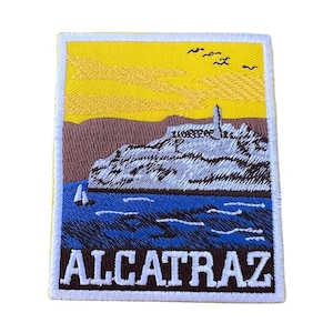 Puede incluir: Un parche bordado con fondo amarillo, que representa una isla gris con un faro, un velero en agua azul, y el texto "ALCATRAZ" en marrón con contornos blancos.