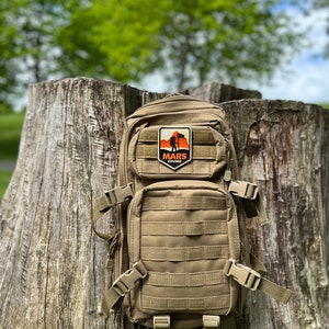 May include: A tan tactical backpack with a patch that says "MARS EXPLORER" and a silhouette of a person standing on a mountain with a flag.