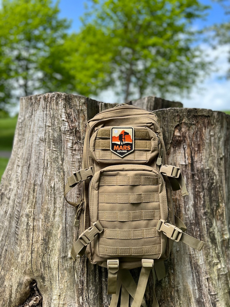 May include: A tan tactical backpack with a patch that says "MARS EXPLORER" and a silhouette of a person standing on a mountain with a flag.