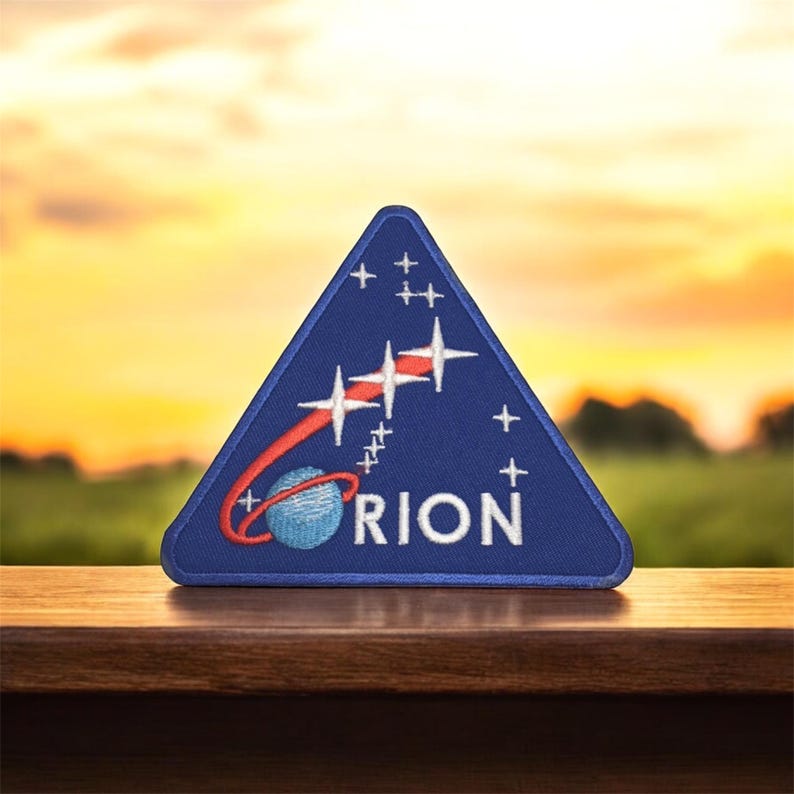 May include: A blue triangular patch with a white embroidered rocket ship and stars. The text "ORION" is embroidered in white below the rocket ship.