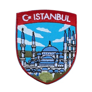 Op de afbeelding: Geborduurde patch in schildvorm met de tekst "C* ISTANBUL" in wit op een rode achtergrond. Het toont een blauwe moskee met witte minaretten tegen een blauwe lucht met wolken. Een reissouvenir.