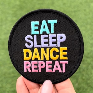 Könnte beinhalten: Ein runder, schwarzer Aufnäher mit dem gestickten Text "EAT SLEEP DANCE REPEAT" in Türkis, Lavendel, Gelb und Rosa. Der Aufnäher wird vor einem grünen Hintergrund gehalten.