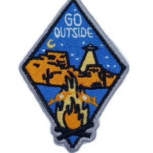 May include: A diamond-shaped embroidered patch with a blue background and the text "GO OUTSIDE" in white. The patch features a campfire, a mountain range, a crescent moon, and a UFO in the night sky.