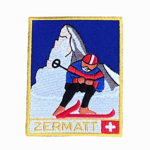 Peut inclure: Patch brodé représentant un skieur en pull rouge et bleu descendant une montagne avec un sommet blanc. Le patch a un fond bleu et une bordure jaune. Le texte "ZERMATT" et un drapeau suisse se trouvent en bas du patch.