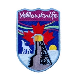 Op de afbeelding: Een blauwe en witte geborduurde patch met de tekst "Yellowknife" in witte letters. De patch toont een rood esdoornblad, een witte wolf, een grijze olieboringstoren en een gele zon.