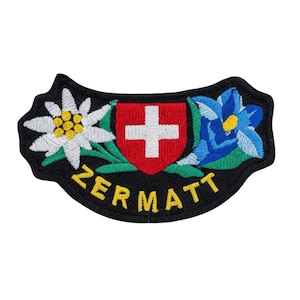 Puede incluir: Parche bordado con un escudo rojo y una cruz blanca, flanqueado por una flor blanca y amarilla y una flor azul. La palabra "ZERMATT" está bordada en amarillo sobre un fondo negro.